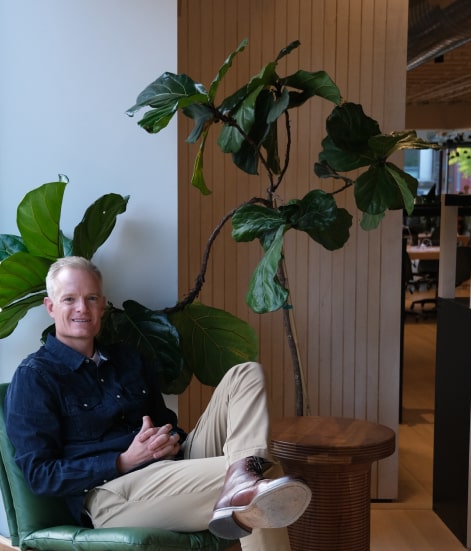 A picture of Jason Kiln in a smart casual attire with a plant background in an office setting.